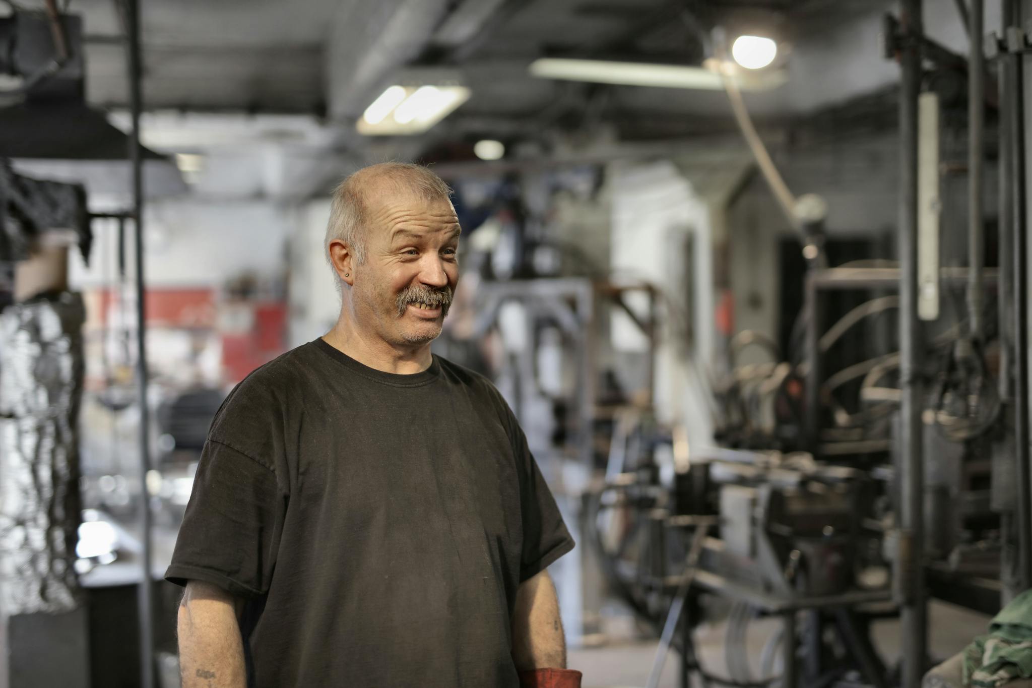 A mature male factory worker smiles while standing in an industrial environment indoors.