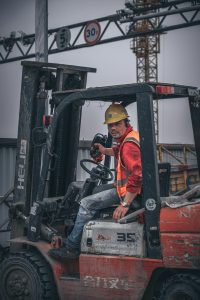 A construction worker wearing a safety helmet operates a forklift on-site under a crane.