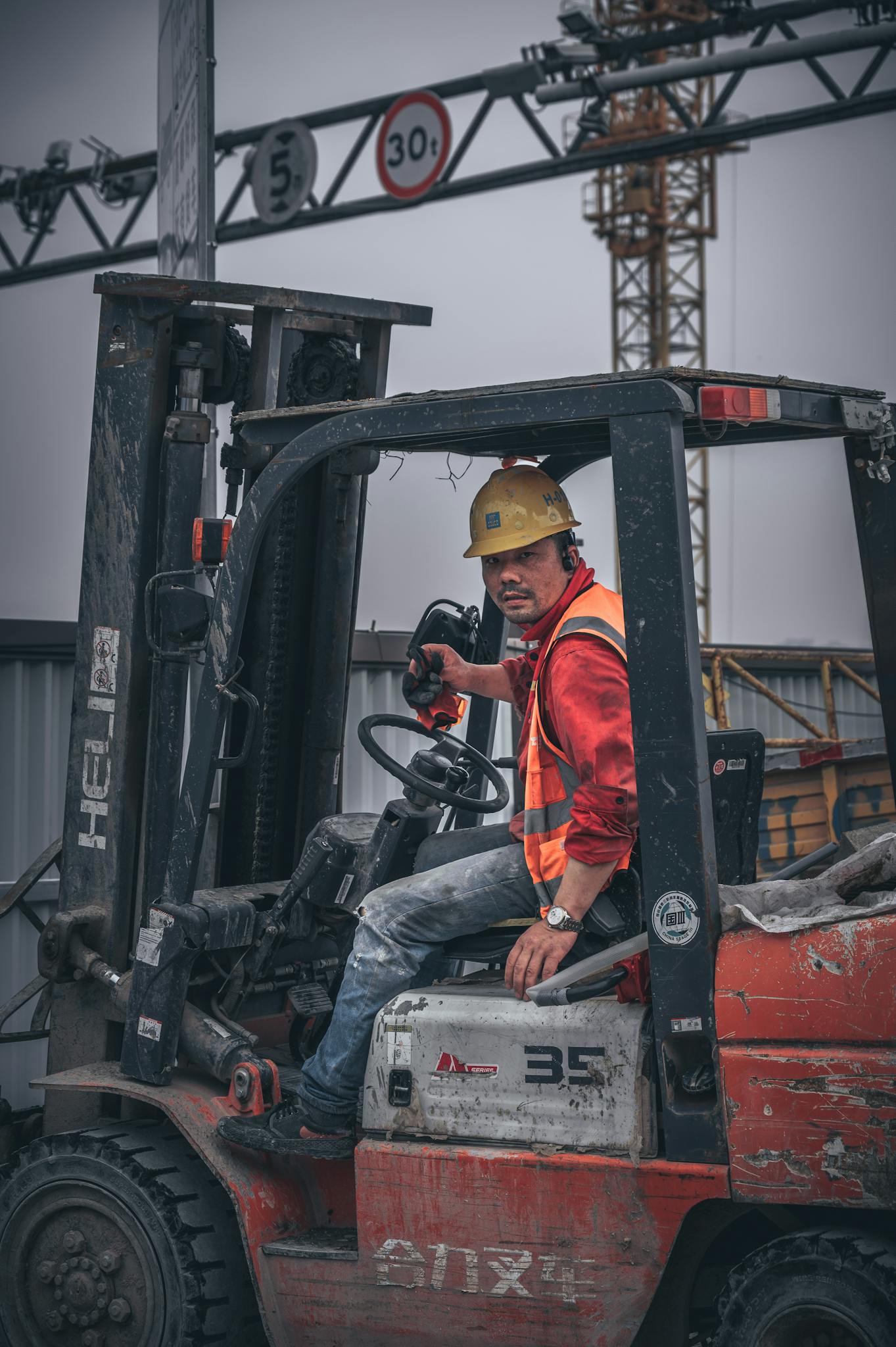 A construction worker wearing a safety helmet operates a forklift on-site under a crane.